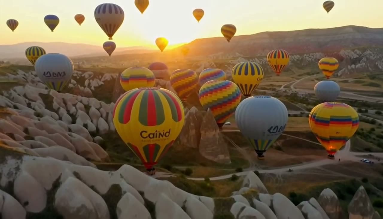 Kapadokya'da gün doğumunda gökyüzünü dolduran sıcak hava balonları, kamera yavaşça yükseliyor. MiniMax H3 Max ile üretilen videodan bir kare.